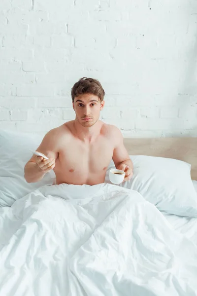 Смущенный человек держит чашку кофе и смартфон в постели — стоковое фото