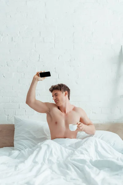 Homme mécontent tenant tasse de café et smartphone avec écran blanc au lit — Photo de stock