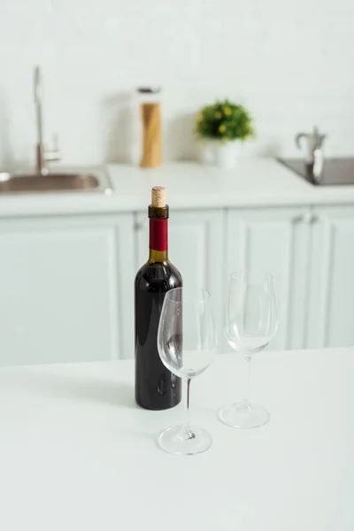 Bottiglia con vino rosso vicino bicchieri vuoti sul tavolo da cucina bianco — Foto stock