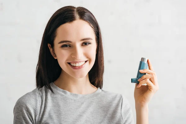 Hermosa chica sonriendo a la cámara mientras sostiene el inhalador sobre fondo blanco - foto de stock