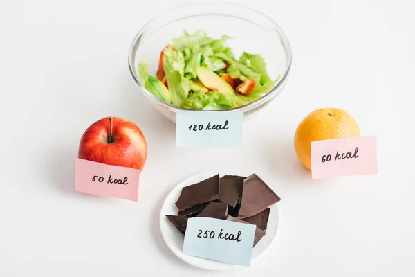 Свежие фрукты, шоколад и салат с калориями на карточках на белом фоне, диета подсчета калорий — стоковое фото