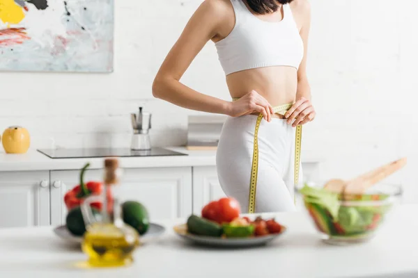 Foco seletivo da mulher magra que mede a cintura com fita perto de verduras frescas e salada na mesa — Fotografia de Stock