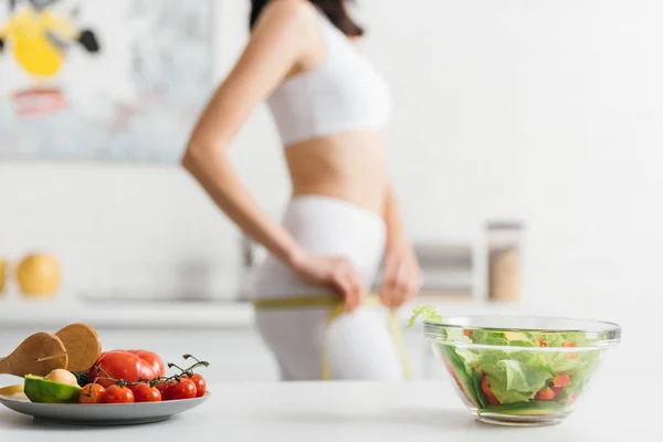 Foco seletivo de verduras orgânicas com salada na mesa e esportista medindo quadris com fita na cozinha — Fotografia de Stock