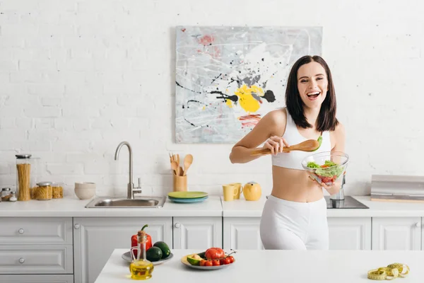 Schöne Sportlerin lächelt in die Kamera, während sie Schüssel mit Salat in der Nähe von reifem Gemüse und Maßband auf dem Küchentisch hält — Stockfoto