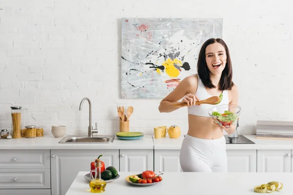 Bella sportiva sorridente alla macchina fotografica mentre tiene la ciotola con insalata vicino a verdure mature e nastro di misurazione sul tavolo della cucina — Foto stock