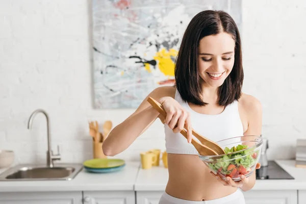 Attractive fit sportswoman smiling while mixing salad on kitchen — Stock Photo