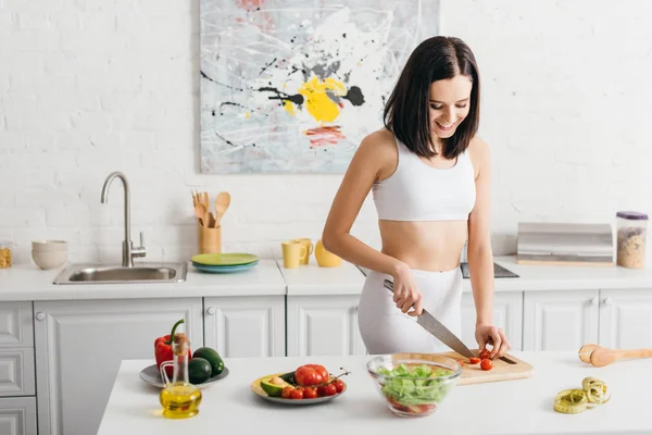 Attraktive Frau lächelt beim Schneiden von Biogemüse in der Nähe von Schüssel mit Salat und Maßband auf Küchentisch — Stockfoto