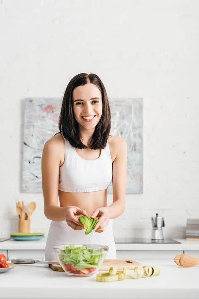 Focus selettivo della sportiva che sorride alla macchina fotografica mentre cucina insalata con verdure fresche e avocado in cucina — Foto stock