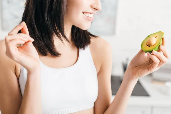 Cropped view of smiling sportswoman holding piece of avocado in kitchen — Stock Photo