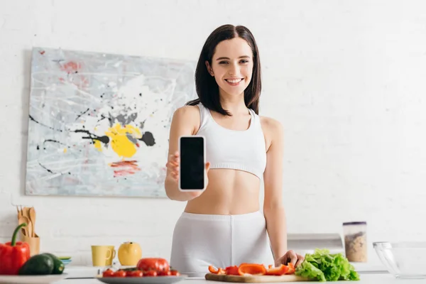 Hermosa deportista mostrando teléfono inteligente y sonriendo a la cámara mientras cocina ensalada fresca en la cocina - foto de stock