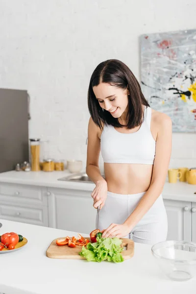 Attractive slim sportswoman smiling and cooking salad with fresh vegetables — Stock Photo