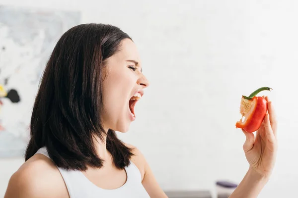 Beautiful girl grimacing while holding piece of bell pepper in kitchen — Stock Photo