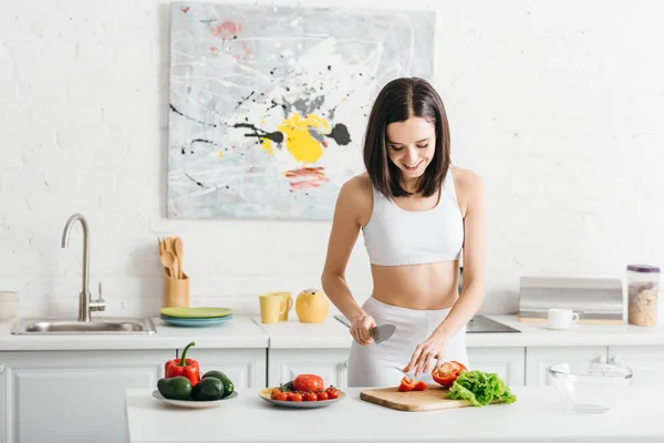 Smiling sportswoman cooking salad with ripe vegetables on kitchen table — Stock Photo