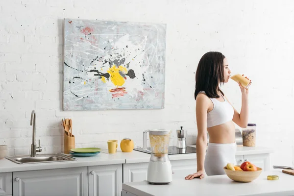 Side view of beautiful slim sportswoman drinking smoothie in kitchen — Stock Photo