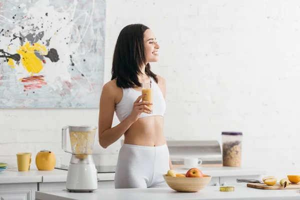 Attractive fit sportswoman smiling and holding glass of smoothie near measuring tape on kitchen table — Stock Photo