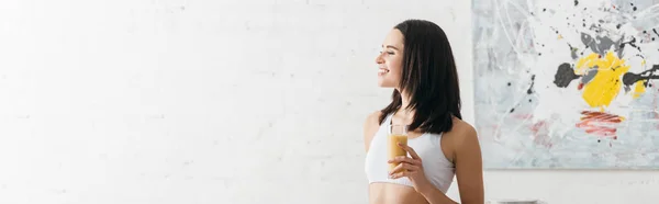 Panoramic shot of beautiful smiling sportswoman holding glass of smoothie at home — Stock Photo