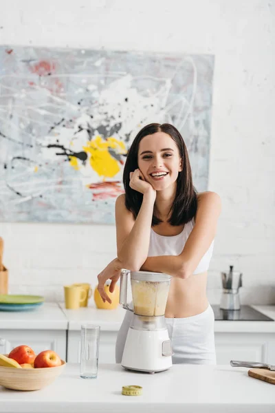 Atractiva deportista en forma preparando batido y sonriendo a la cámara cerca de cinta métrica en la mesa de la cocina - foto de stock