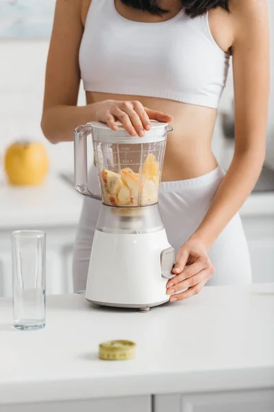 Cropped view of fit sportswoman preparing smoothie with fresh fruits near measuring tape on kitchen table — Stock Photo