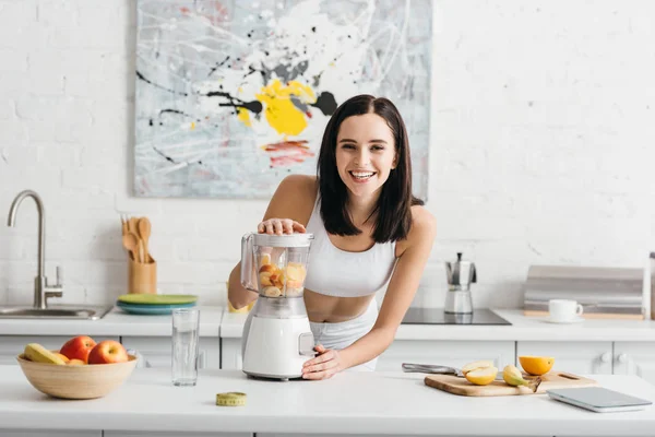 Selective focus of smiling sportswoman preparing smoothie near measuring tape and fruits on kitchen table, calorie counting diet — Stock Photo