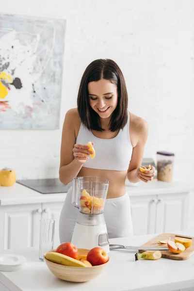 Усміхнена спортсменка додає свіжі фрукти в блендері на кухонному столі — стокове фото