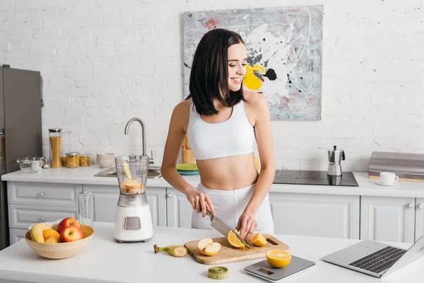 Lächelnde fitte Sportlerin, die auf Laptop schaut, während sie Obst in der Nähe des Mixers auf dem Küchentisch schneidet, Kalorienzähler-Diät — Stockfoto