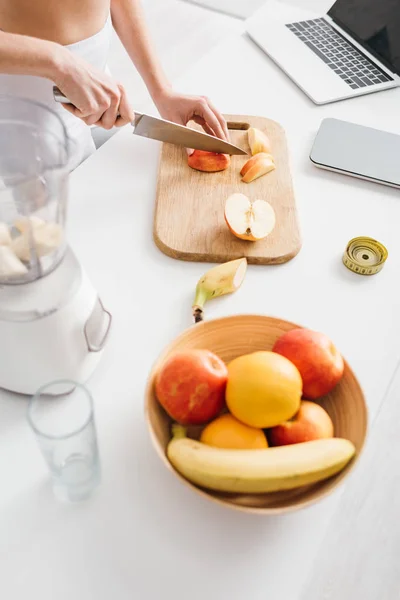 Visão de alto ângulo da mulher cortando frutas para smoothie com fita métrica, balanças e laptop na mesa da cozinha, dieta de contagem de calorias — Fotografia de Stock