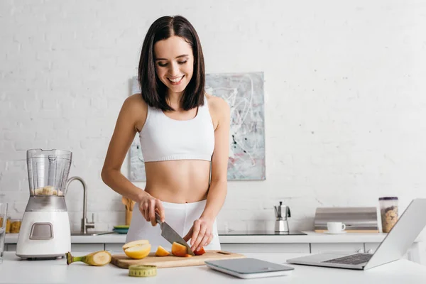 Atractiva deportista sonriendo mientras corta frutas para batido cerca de la computadora portátil, escalas y cinta métrica en la mesa, dieta de conteo de calorías - foto de stock