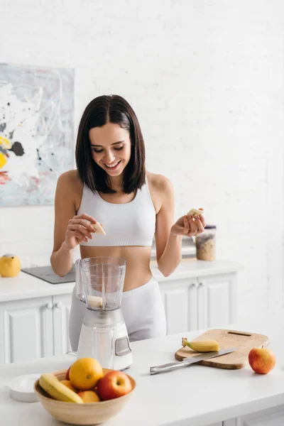 Hermosa deportista sonriente poniendo plátano en la licuadora mientras prepara batido - foto de stock