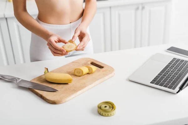 Vue recadrée d'une femme en forme coupant la banane près d'un ordinateur portable, d'un smartphone et d'un ruban à mesurer sur la table — Photo de stock