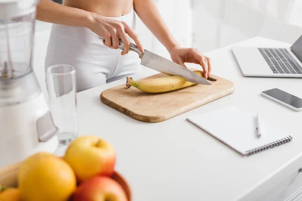 Abgeschnittene Ansicht eines fitten Mädchens beim Schneiden von Bananen in der Nähe digitaler Geräte und Notizbücher auf dem Tisch — Stockfoto