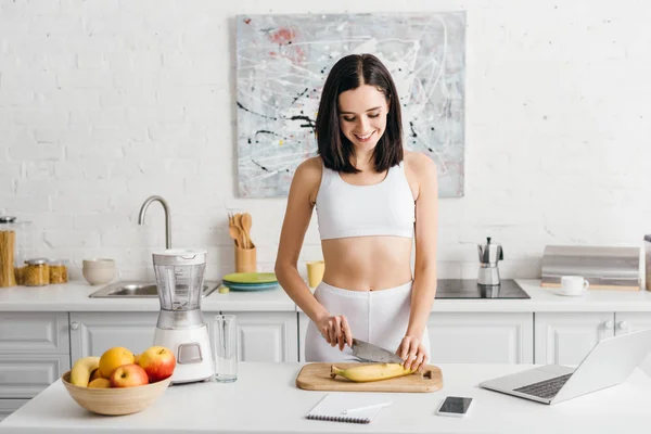 Femme sportive souriante préparant un smoothie près d'un ordinateur portable, d'un smartphone et d'un ordinateur portable sur la table dans la cuisine — Photo de stock