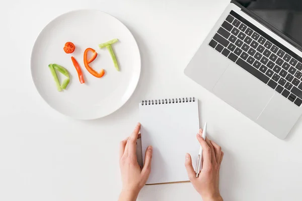 Vista superior de la mujer que sostiene la pluma y el cuaderno cerca de la dieta de letras de rebanadas de verduras en el plato y el ordenador portátil en el fondo blanco - foto de stock