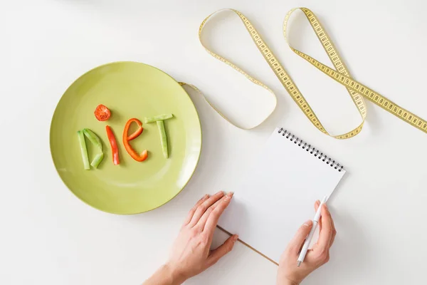 Visão superior da mulher escrevendo em notebook perto de fita métrica e placa com letras de dieta de fatias de vegetais no fundo branco — Fotografia de Stock