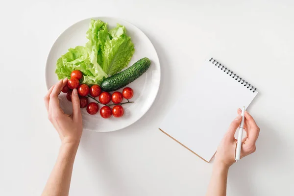 Vista superior de la mujer que sostiene la pluma cerca del cuaderno y verduras frescas en el plato sobre fondo blanco - foto de stock