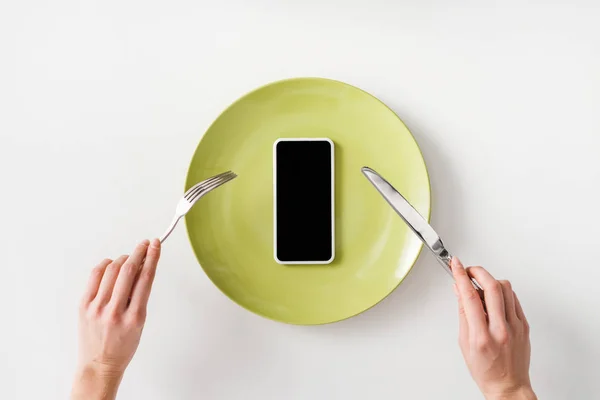 Vista superior de la mujer que sostiene los cubiertos cerca del teléfono inteligente en la placa sobre fondo blanco - foto de stock