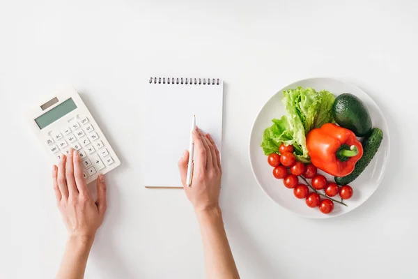 Visão superior da mulher usando calculadora perto de notebook e legumes frescos na placa no fundo branco — Fotografia de Stock