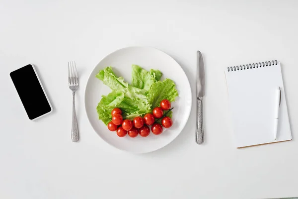 Вид сверху свежих овощей на тарелке, смартфоне и ноутбуке на белом фоне — стоковое фото