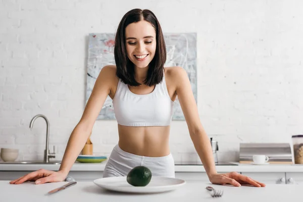 Lächelnde Sportlerin mit Blick auf Avocado auf Teller auf Tisch — Stockfoto