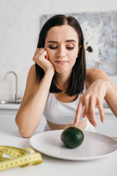 Selective focus of thoughtful sportswoman looking at avocado near measuring tape on kitchen table — Stock Photo