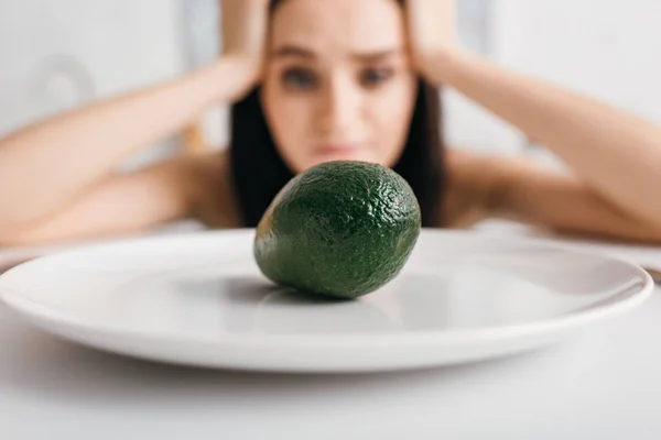 Вибірковий фокус стомлюючої дівчини, дивлячись на авокадо на тарілці на столі — стокове фото
