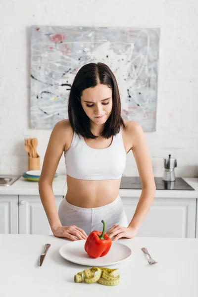 Thoughtful sportswoman looking at bell pepper near measuring tape on kitchen table — Stock Photo