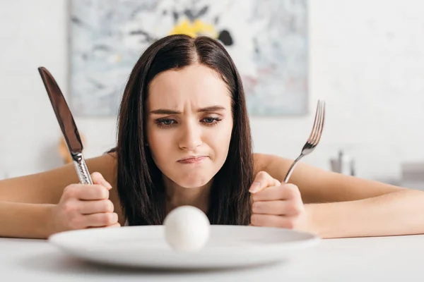 Foco seletivo de menina pensativa segurando talheres e olhando para o ovo na placa na mesa da cozinha — Fotografia de Stock