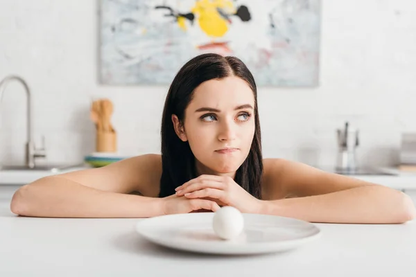 Chica pensativa mirando lejos cerca de huevo en plato en la mesa de la cocina - foto de stock
