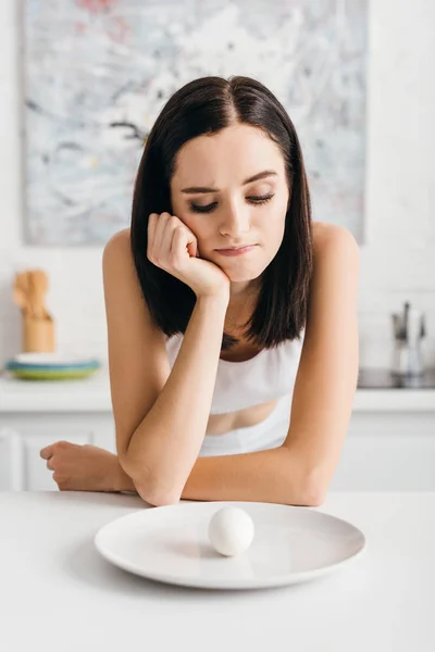 Pensive young sportswoman looking at egg on plate in kitchen — Stock Photo