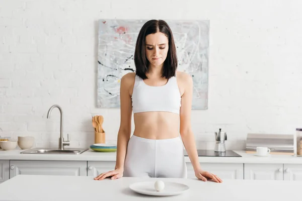 Sportive réfléchie regardant oeuf sur la plaque sur la table de cuisine — Photo de stock