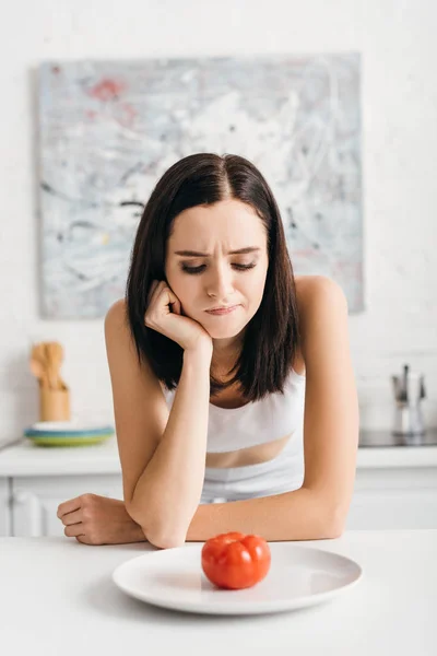 Pensive sportswoman looking at ripe tomato on plate on table — Stock Photo