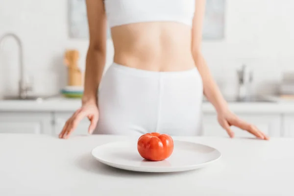Selective focus of ripe tomato on plate and slim sportswoman near kitchen table — Stock Photo