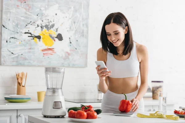 Desportista sorrindo usando smartphone perto de escalas, legumes e fita métrica na mesa da cozinha, dieta de contagem de calorias — Fotografia de Stock