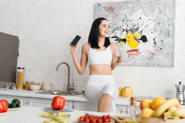 Focus selettivo di sportiva sorridente con bicchiere d'acqua e smartphone vicino a verdure, frutta e nastro di misurazione sul tavolo da cucina, dieta per il conteggio delle calorie — Foto stock