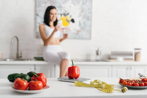 Selective focus of fresh vegetables with measuring tape near scales and sportswoman with smartphone and glass of water in kitchen, calorie counting diet — Stock Photo